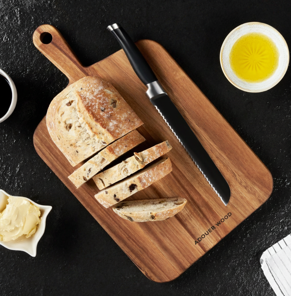 Wooden cutting board with sliced bread, a knife, and a small bowl of oil on a dark surface.