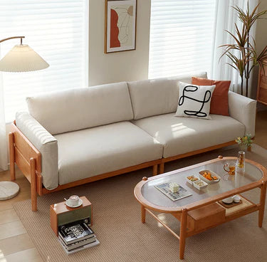 A cream upholstered cane sofa with wooden frame, placed in a well-lit living room setting, accompanied by a glass coffee table with items on it and a lamp in the background.