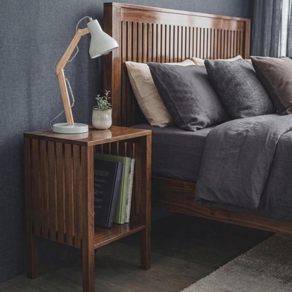 Wooden nightstand with lamp and books next to a bed with pillows.