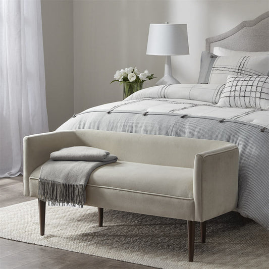 Beige bench in front of a bed with gray bedding in a bedroom setting.