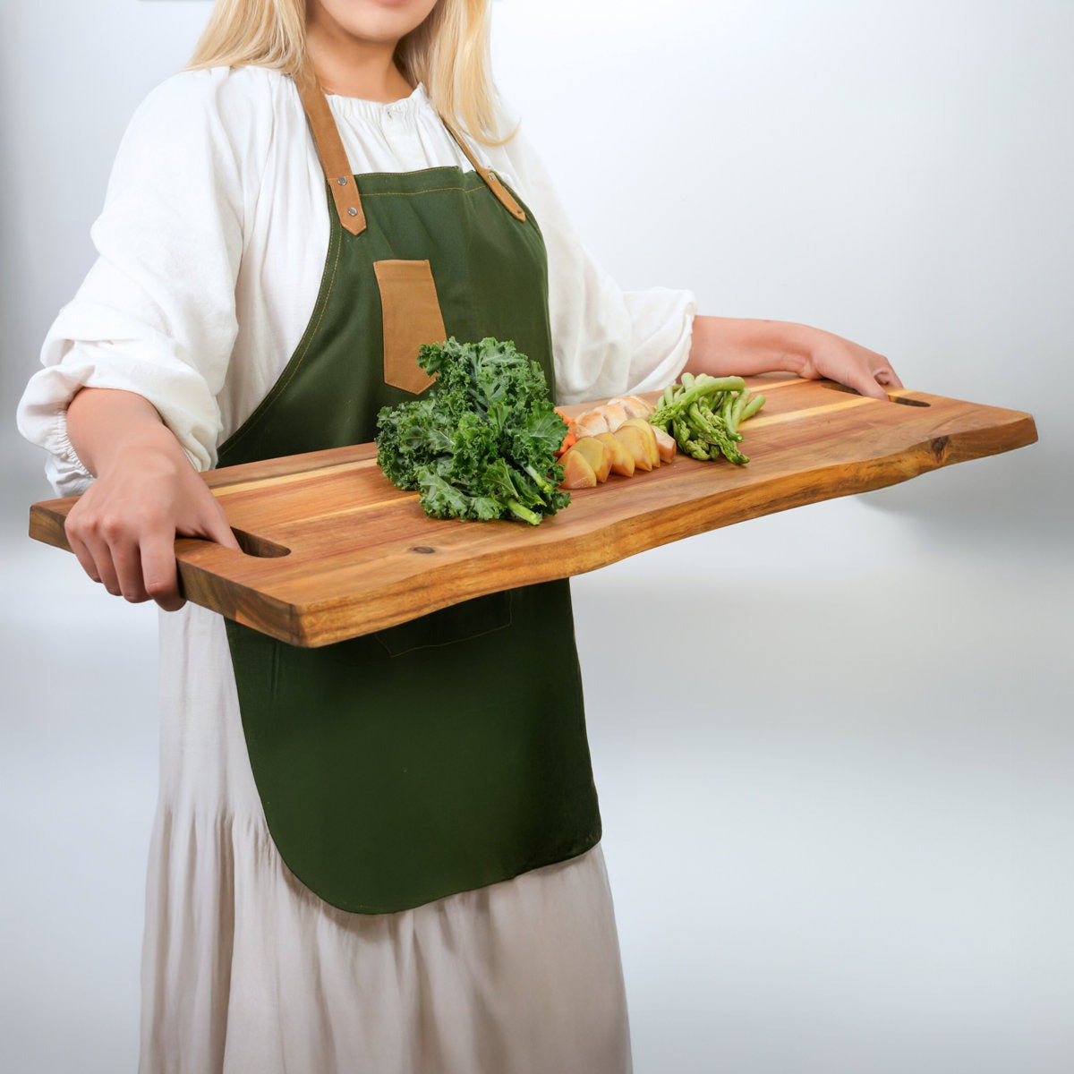 Person holding a wooden cutting board with vegetables wearing a green apron on a plain background