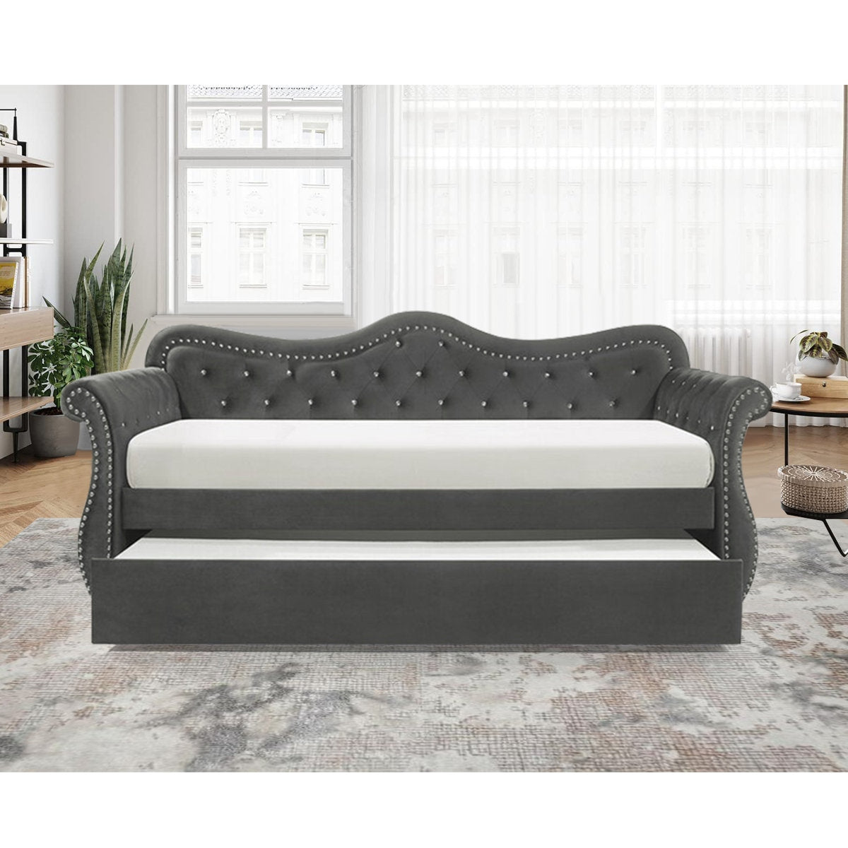 Gray upholstered bed with white mattress in a room with large windows and plants.