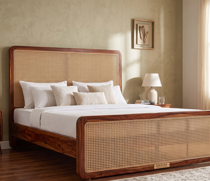 A wooden bed frame with woven cane panels on the headboard and footboard, styled in a warm, neutral-toned bedroom with white bedding, a bedside lamp, and a textured beige wall.