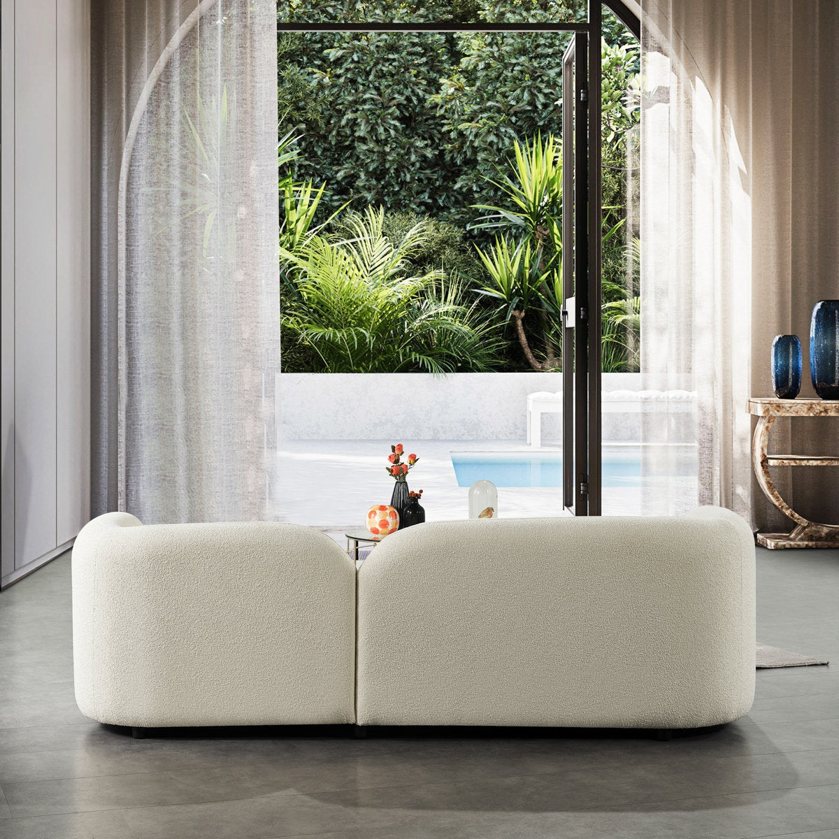 Modern white sofa in a room with large windows showing a garden and pool.