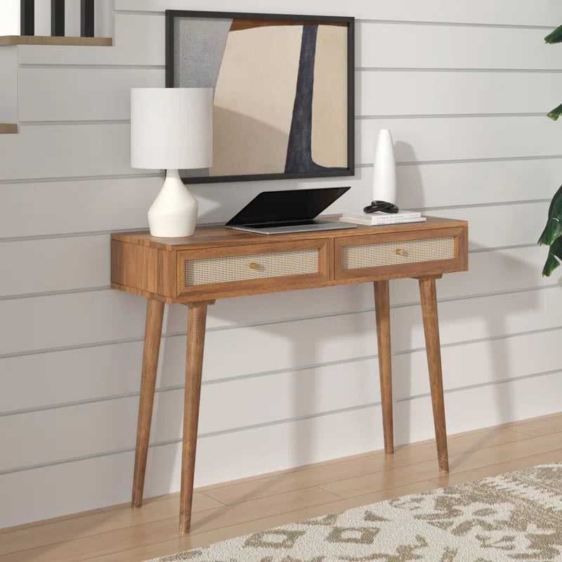 A wooden console table with drawers, featuring a lamp and a framed picture on top, set against a white panelled background.