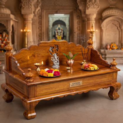 A large carved wooden altar table in a temple-like interior, decorated with a small Ganesh statue, lit oil lamps, incense, and fruit plates. The front has a brass nameplate reading 'ADOUER'.