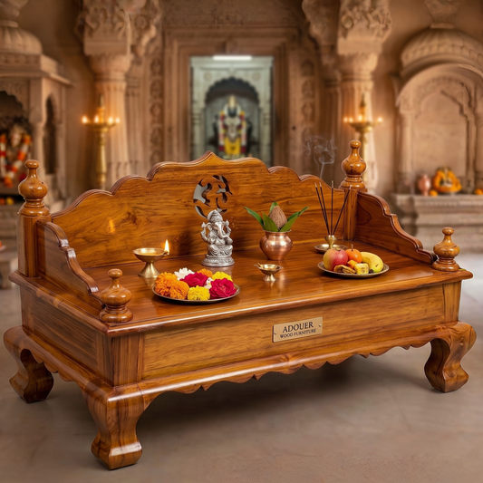 A large carved wooden altar table in a temple-like interior, decorated with a small Ganesh statue, lit oil lamps, incense, and fruit plates. The front has a brass nameplate reading 'ADOUER'.