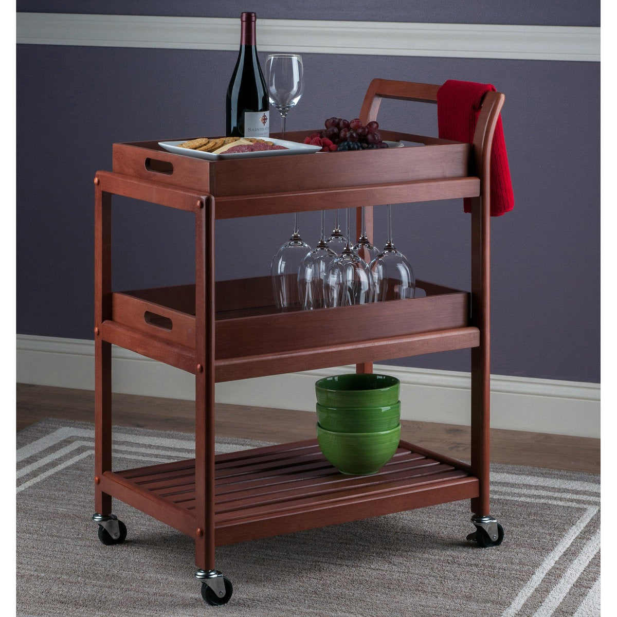 Wooden serving cart with wine bottle, glasses, and food on a gray carpeted floor.