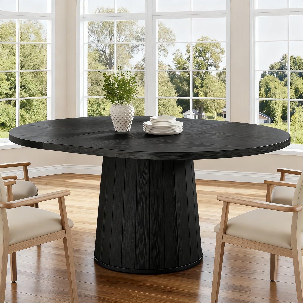 Round black dining table with chairs in a room with large windows showing greenery outside.