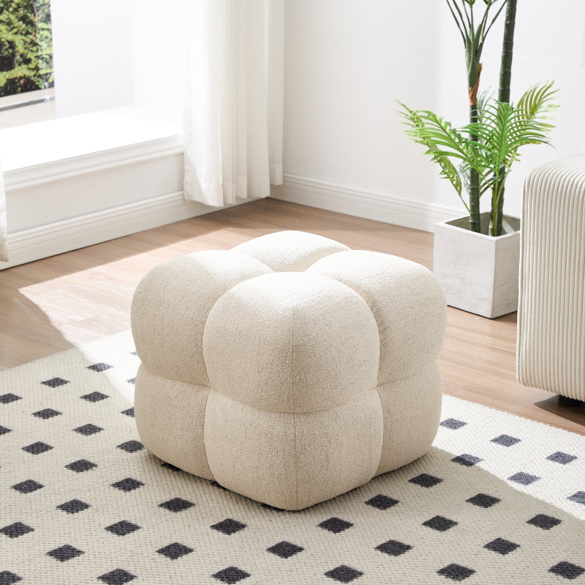 Beige cube-shaped ottoman in a room with a patterned rug, plant, and window.