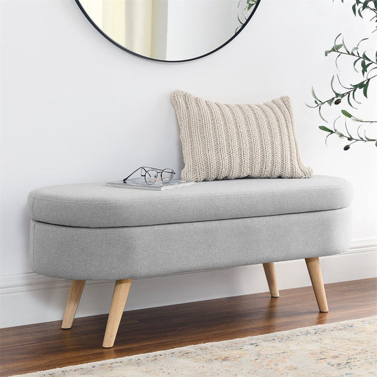 Gray upholstered bench with wooden legs against a white wall, featuring a pillow and glasses on top.