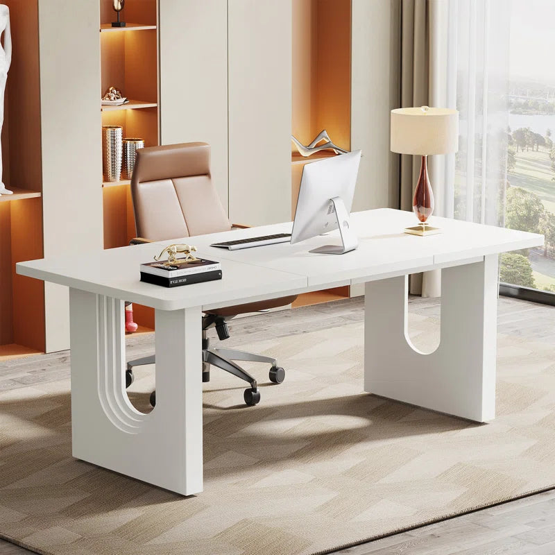 A sleek white executive desk with sculpted panel legs in an office setting, with an office chair and lamp in the background.