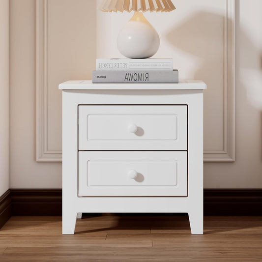 A white wooden bedside table with two drawers and knob handles, placed on a hardwood floor against a white wall, featuring a lamp and a book on top.