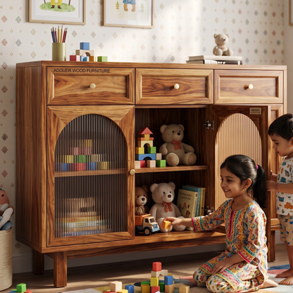 A wooden children's storage cabinet with glass-front doors and open shelves, styled in a child's room with toys inside. A girl in patterned pajamas sits on the floor beside it, and part of a boy is visible at the right edge.