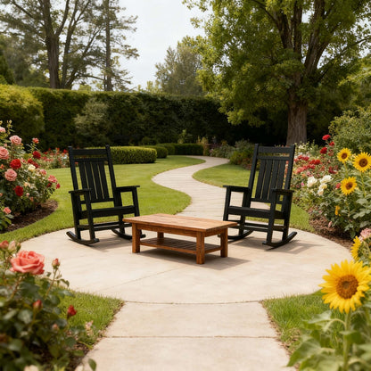 Two black rocking chairs with a wooden table on a garden path surrounded by flowers and greenery.