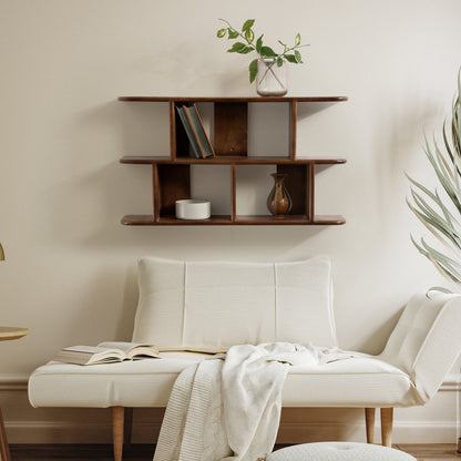 White sofa with a book and blanket, wooden shelf above with decor items, and plant in the corner.