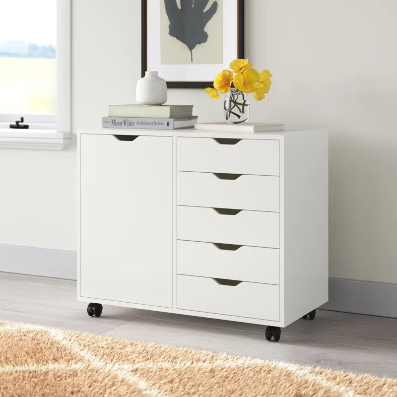 White wooden file cabinet with four drawers and a cabinet door, set on casters and positioned against a wall with a framed picture above it, a vase with yellow flowers on top, and some books beside it.