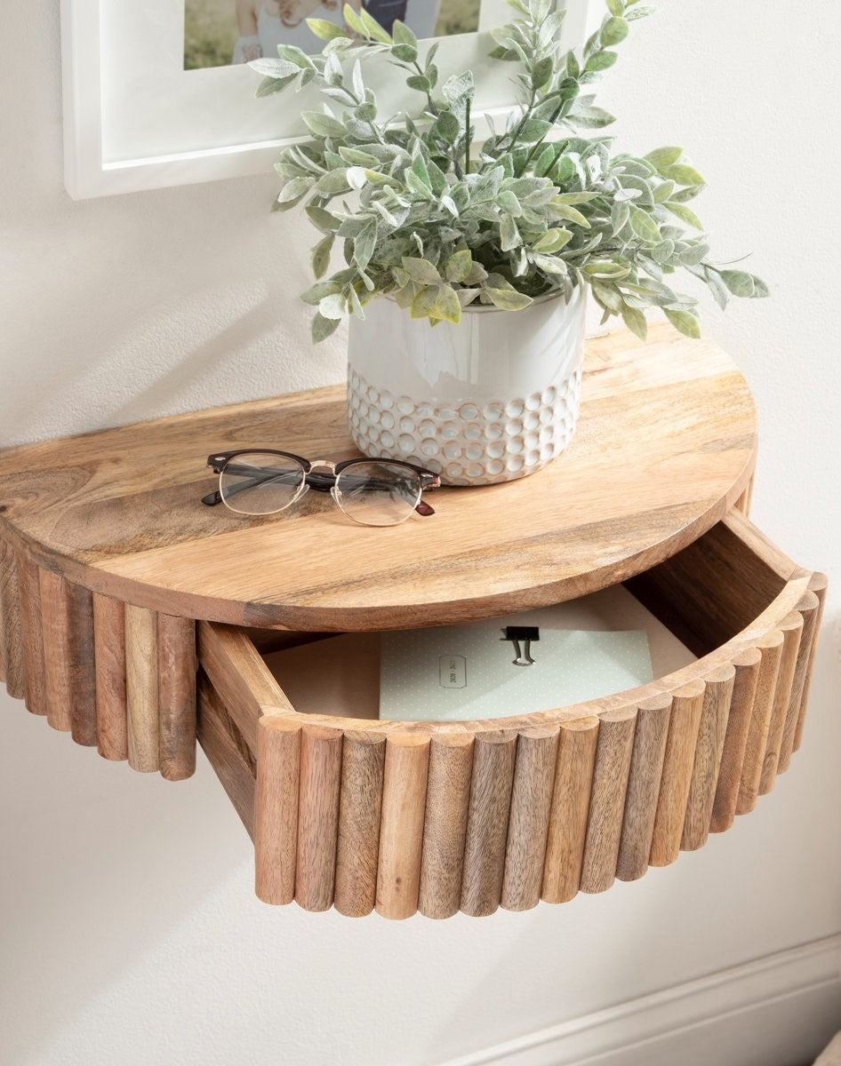Wooden wall-mounted shelf with a plant, glasses, and a box on a white wall.