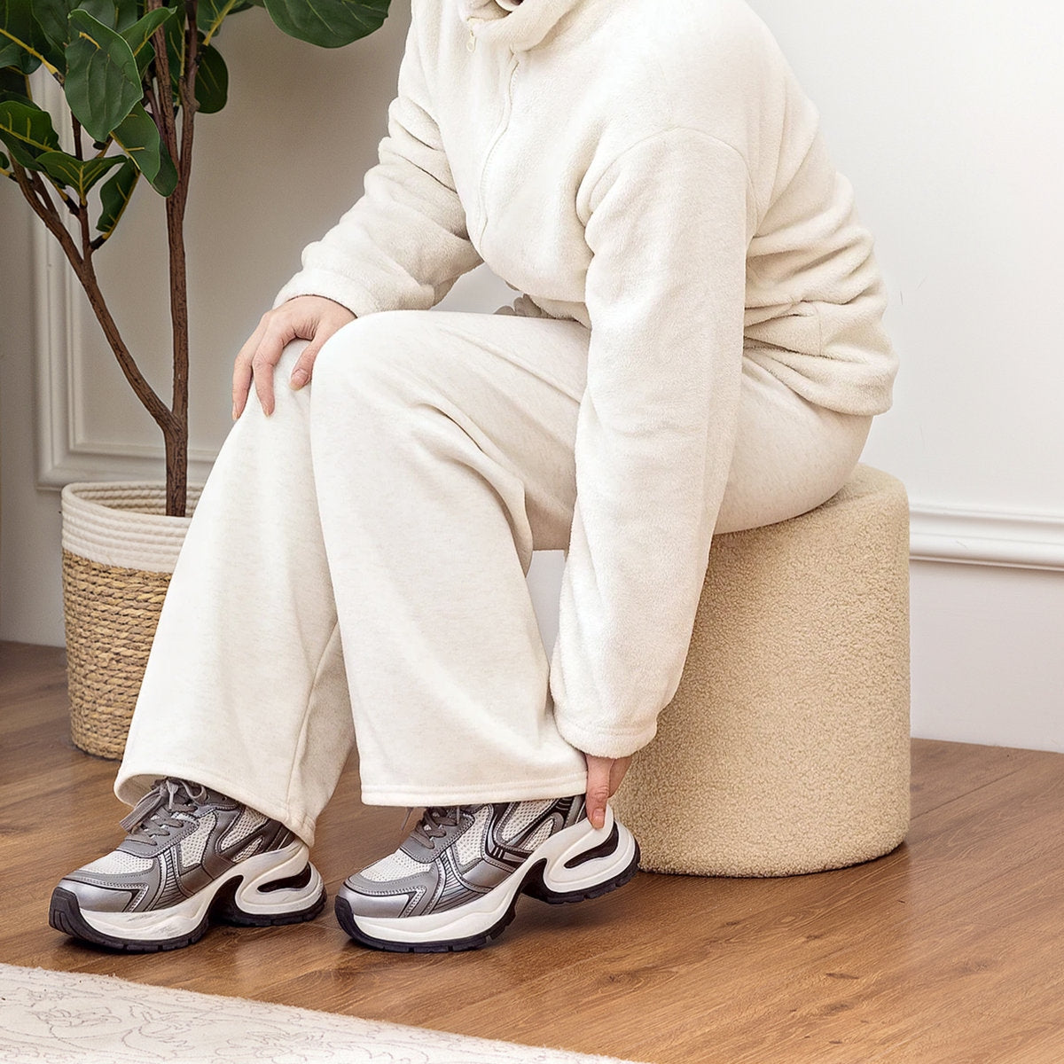 Person wearing a white tracksuit and sneakers sitting on a beige stool indoors.