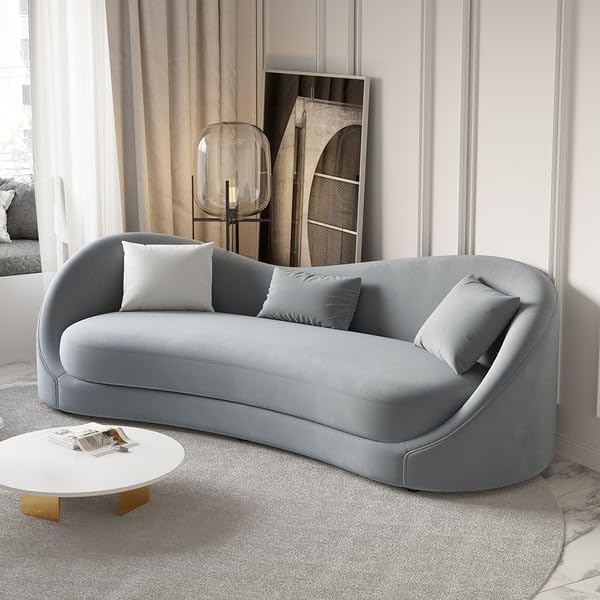 An emerald green modern curved sofa with three cushions, placed in a well-lit room with a white curtain and a decorative wall panel behind it. The sofa is accompanied by two white round coffee tables with a gold accent on the larger table.