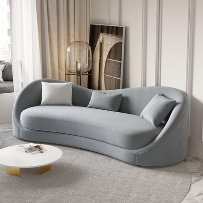 An emerald green modern curved sofa with three cushions, placed in a well-lit room with a white curtain and a decorative wall panel behind it. The sofa is accompanied by two white round coffee tables with a gold accent on the larger table.