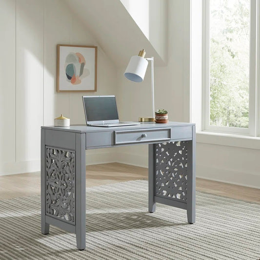 A gray Sheesham wood study table with carvings on the legs, featuring a drawer for storage, a laptop, a lamp, and a notebook placed on the table, set in a room with a light interior.