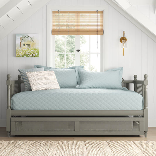 A gray and white Sheesham wood trundle bed with a mattress and bedding, placed in a room with a window and a decorative picture on the wall.