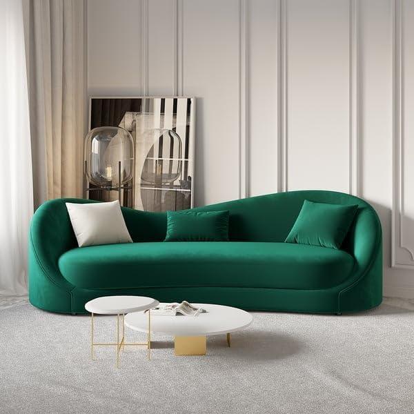 An emerald green modern curved sofa with three cushions, placed in a well-lit room with a white curtain and a decorative wall panel behind it. The sofa is accompanied by two white round coffee tables with a gold accent on the larger table.