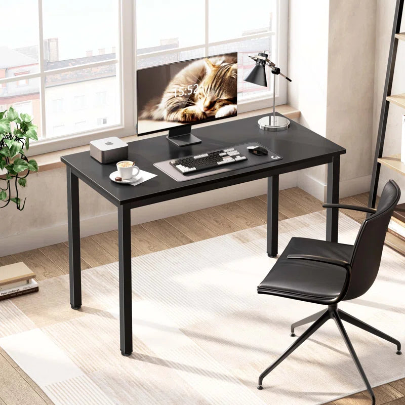 A modern black computer desk with a black office chair, set in an office environment with decorative items and a potted plant. The desk has a wooden tabletop and metal frame.