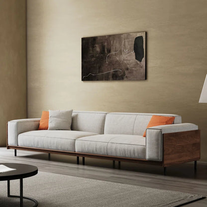 A modern sofa with square arms, solid wood frame, and fabric upholstery, featuring beige and orange cushions, positioned in a well-lit room with a decorative painting above.