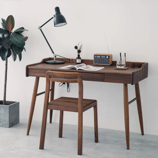 A modern wooden office desk made of Sheesham wood with a walnut finish, accompanied by a matching chair, lamp, and decorative items. The desk has a drawer and is set in a home workspace with a potted plant and a radio on top.