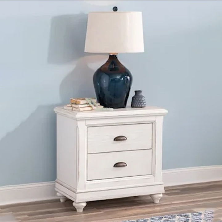 A rustic white two-drawer bedside table made of Sheesham wood, with a lamp and decorative items on top, set against a blue wall.
