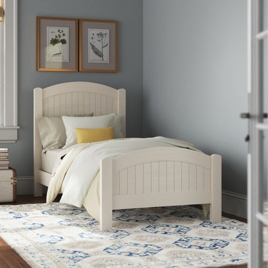 A white finish solid sheesham wood classic bed for kids, with a mattress and pillows, placed in a bedroom setting with decorative items on the wall and a patterned carpet on the floor.