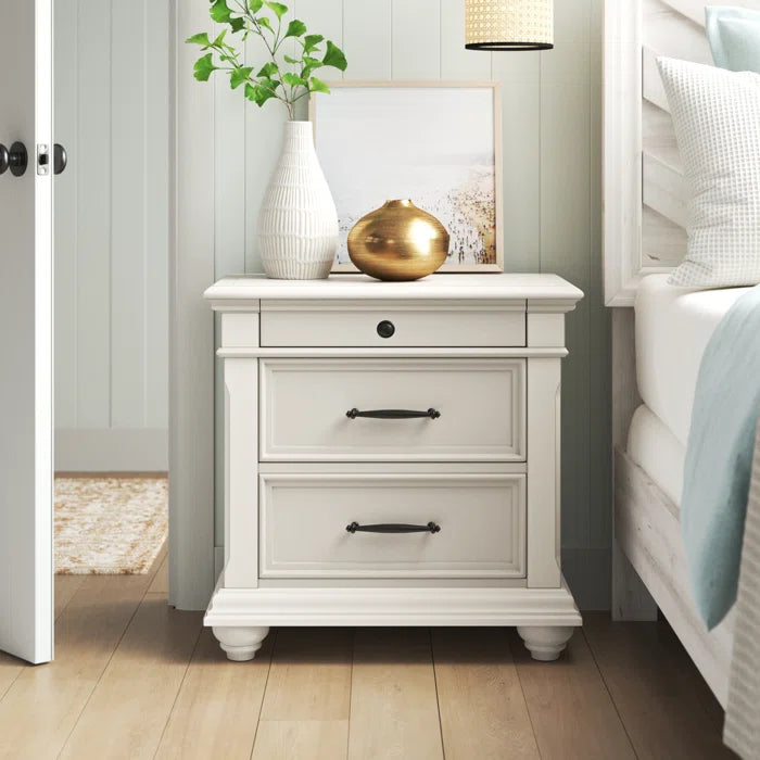 A white wooden bedside table with three drawers, placed in a bedroom setting with a vase and decorative items on top.