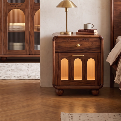 A dark brown wooden nightstand with arched glass-front doors and warm internal lighting, styled in a bedroom setting next to a bed with a brass table lamp on top.