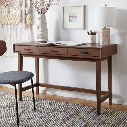 A wooden study and writing desk with three drawers, positioned against a wall with a chair to its left, a framed picture above, and a decorative lamp on the desk.
