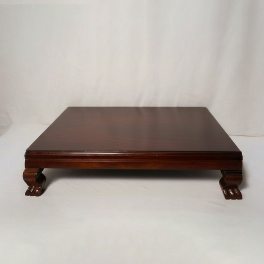 A square dark reddish-brown wooden platform or low table with carved legs, photographed against a white fabric backdrop.