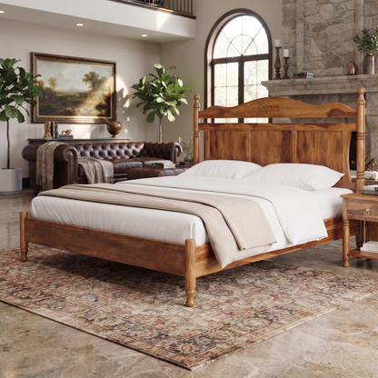 A wooden twin bed with a carved headboard and matching footboard, dressed in white linens and a beige throw, set in a rustic bedroom with a stone fireplace, leather sofa, and patterned rug.