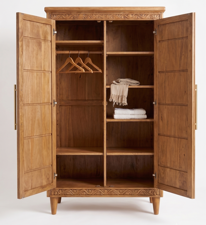 A large carved wooden wardrobe with two doors open, showing interior shelves, hangers, folded textiles, and a scarf. The cabinet has a warm medium-brown wood finish and decorative relief carving along the top and bottom.