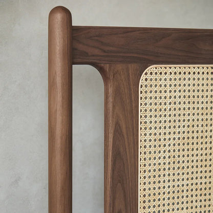 Close-up of a wooden chair or bench showing a dark walnut wood frame with a round finial on the leg and a natural woven cane panel.