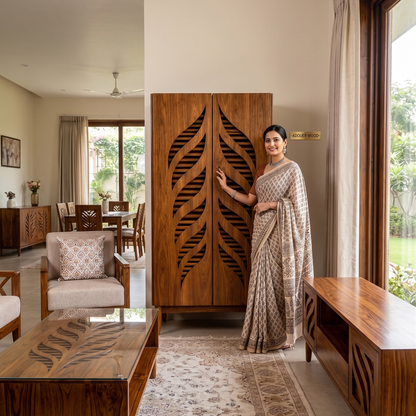 A woman in a beige and brown patterned saree stands next to a large carved wooden wardrobe in a modern, well-lit living room with warm neutral tones.