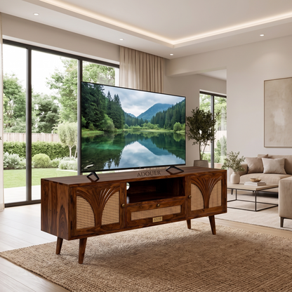 A mid-century modern wooden TV stand with woven cane panels and a central drawer, shown in a bright, contemporary living room with a large television on top. The TV displays a lake landscape, and the room features a beige rug, a sofa, and large windows overlooking a green garden.