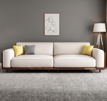 A modern cream leather sofa with a dark wood base, styled with two yellow pillows and one gray patterned pillow, set against a dark gray wall with a minimalist art print and a floor lamp.