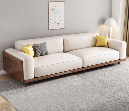 A modern three-seater sofa with cream upholstery and a dark wood base, styled in a minimalist living room with two accent pillows, a gray rug, and a wall-mounted abstract print.