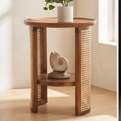 A small round wooden side table with two shelves, featuring woven cane panels on the sides and a decorative shell sculpture on the lower shelf. A white planter sits on the top surface.
