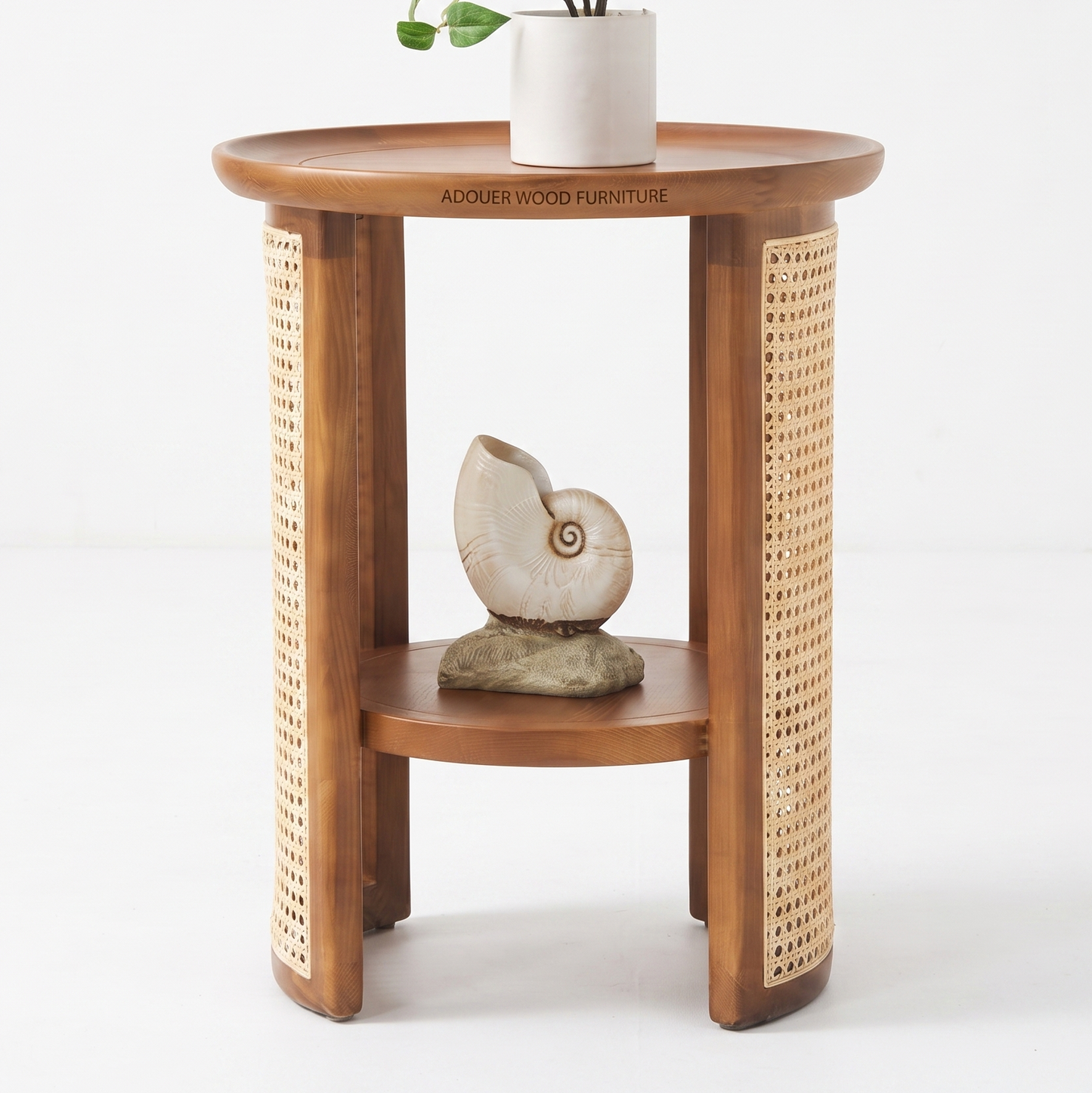 A round wooden side table with a warm medium-brown finish, featuring woven cane panels on the outer sides and a lower shelf. The table is styled with a white ceramic pot and a seashell sculpture, set against a clean white studio background.