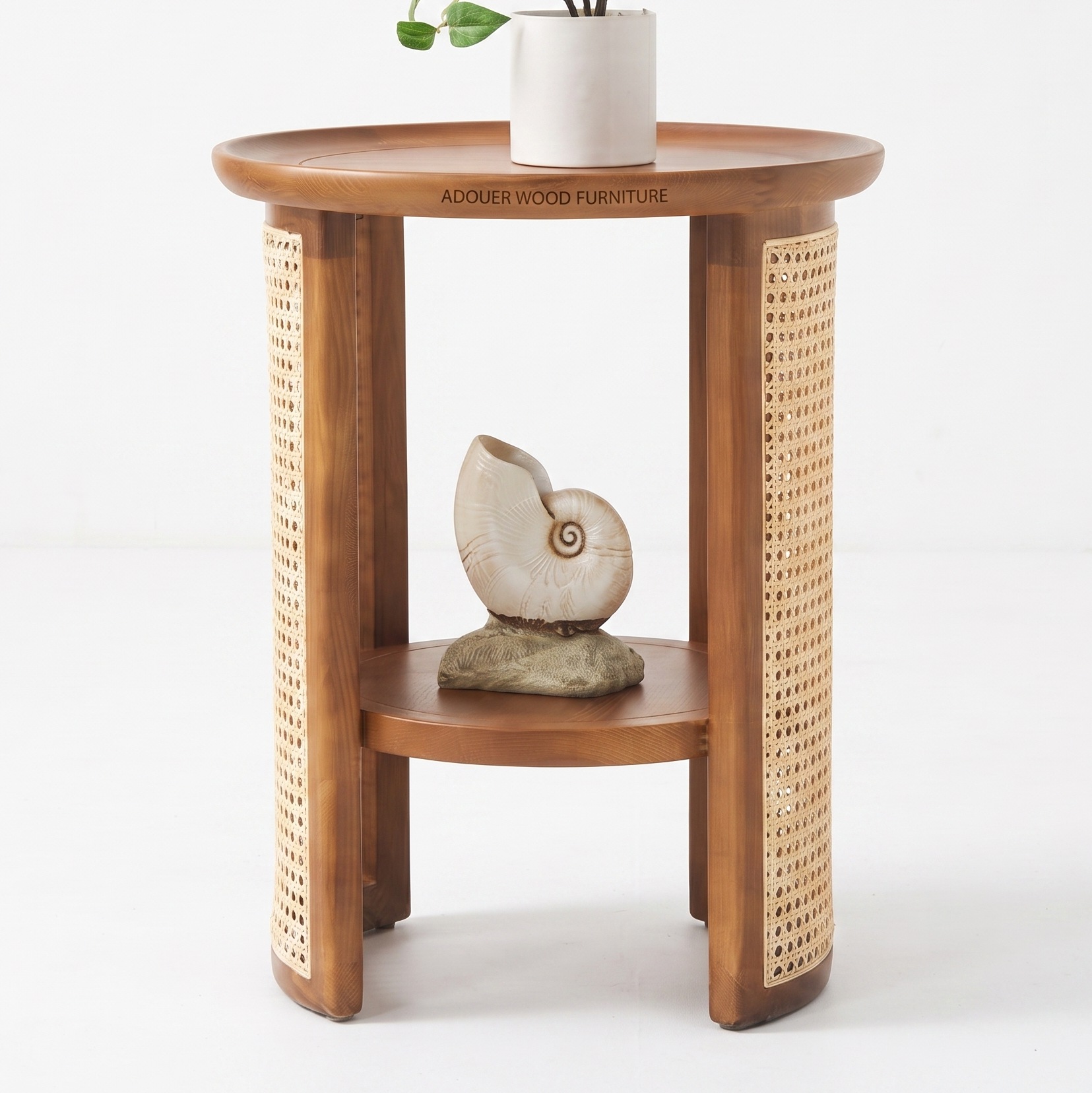 A round wooden side table with a warm medium-brown finish, featuring woven cane panels on the outer sides and a lower shelf. The table is styled with a white ceramic pot and a seashell sculpture, set against a clean white studio background.