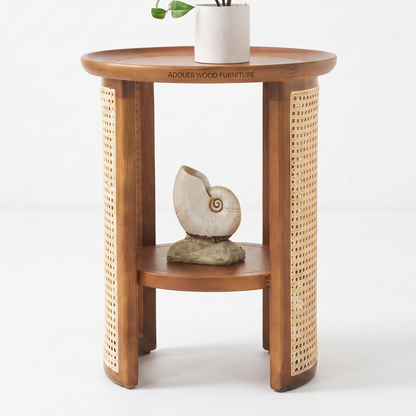 A round wooden side table with a warm medium-brown finish, featuring woven cane panels on the outer sides and a lower shelf. The table is styled with a white ceramic pot and a seashell sculpture, set against a clean white studio background.