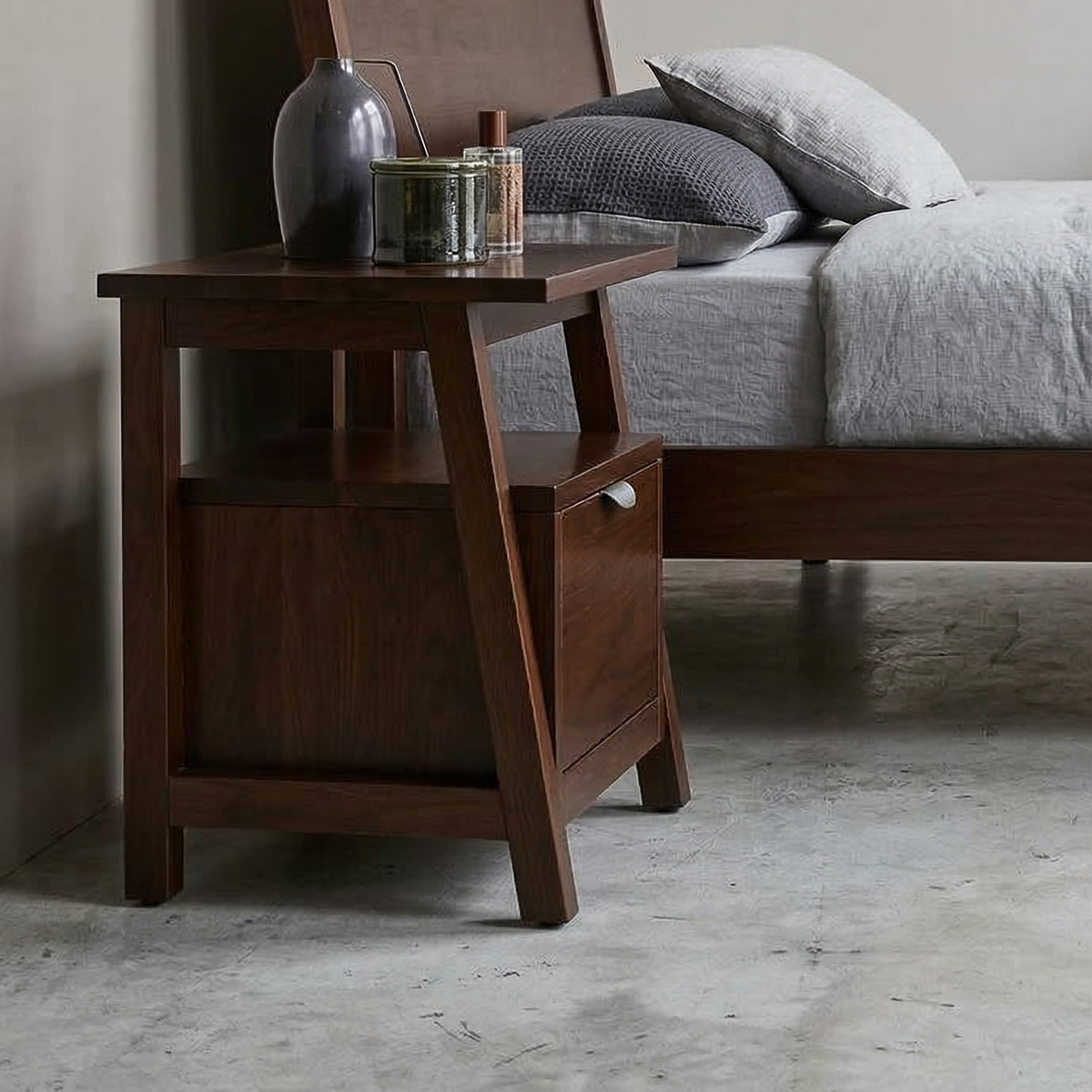 Wooden nightstand with a drawer next to a bed with gray bedding.