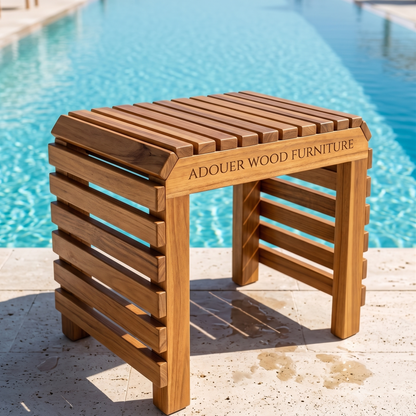 A wooden poolside stool with a slatted top and side panels, engraved with the text 'ADOUER WOOD FURNITURE', set on a stone deck with a swimming pool in the background.